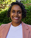 An individual with curly hair, wearing a light pink blazer over a white top, standing outdoors with a green, leafy background.