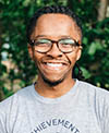 A smiling individual wearing glasses and a light gray T-shirt, standing outdoors with a blurred background of greenery.
