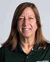 A smiling individual with shoulder-length brown hair, wearing a dark green collared shirt, set against a plain light gray background.