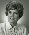 Sheila Fitzgerald smiles for the camera while in front of a neutral background in this black and white photo. Fitzgerald wears a white button-down shirt, with pearl necklace and pearl earrings. Fitsgerald's hair is light colored and short.