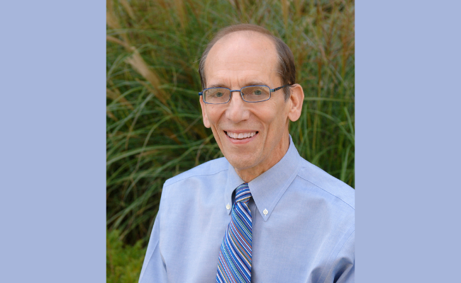 A professional portrait of the same person wearing glasses and a light blue dress shirt with a striped tie, smiling against a natural backdrop of greenery.