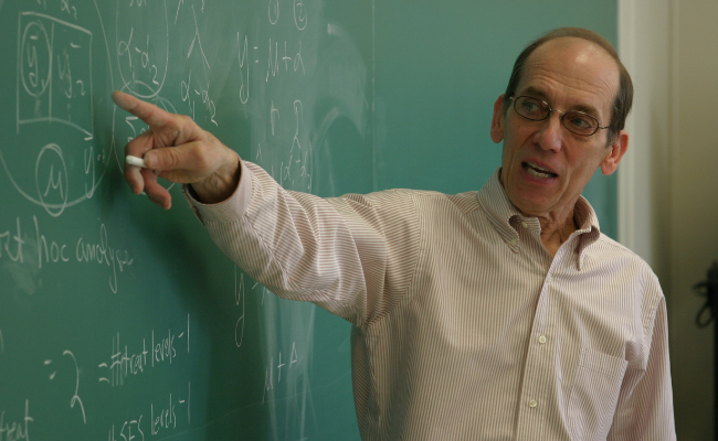 Schmidt wearing glasses and a light-colored dress shirt, gestures toward a chalkboard filled with mathematical equations and diagrams while speaking.