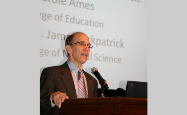 Bill Schmidt wearing glasses and a suit is speaking at a podium with a microphone. The background shows projected text mentioning "College of Education" and "Natural Science."