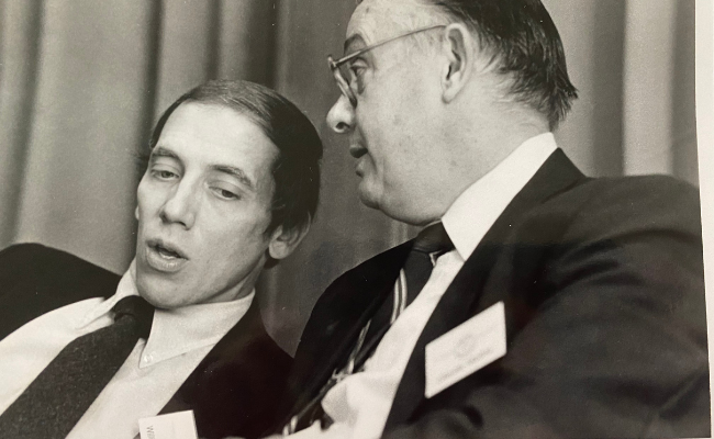 A black-and-white photograph showing a younger William Schmidt engaged in conversation with an older man in formal attire. Schmidt is speaking while the other man listens attentively, possibly during a conference or academic event.