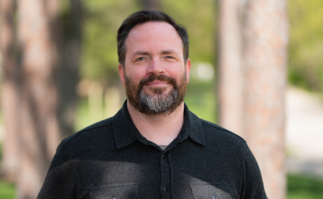 Andrew Driska headshot. Driska wears a black button-down shirt. Driska's dark hair is short. Driska has a beard.