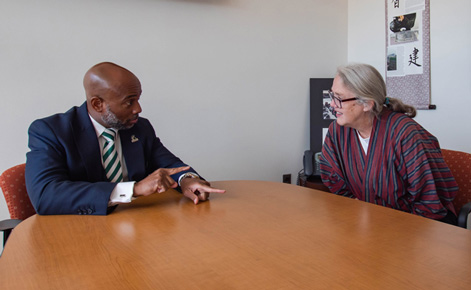  man in a dark suit and striped green-and-white tie (left) sits at a conference table, gesturing with his hand while speaking to a woman on the right. The woman, wearing a red and blue striped cardigan and glasses, listens attentively with a smile. They are seated in an office space with a scroll featuring Asian calligraphy and a framed picture in the background.