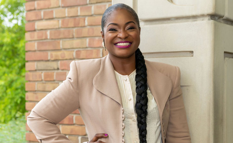 Raven L. Jones headshot. Raven stands near a building outside. Raven wears a beige blazer over a cream-colored blouse. Raven's black hair is tied in a braid. Behind Raven, the building facade is made of reddish brick and concrete. Green leaves are visible in the background. 