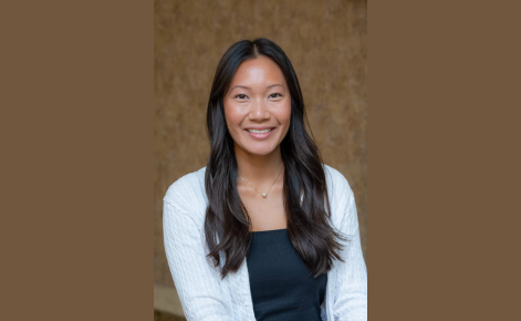 Portrait of Brianna Bui, a young woman with long, dark brown hair wearing a white cardigan over a black top, smiling at the camera against a neutral-toned background."