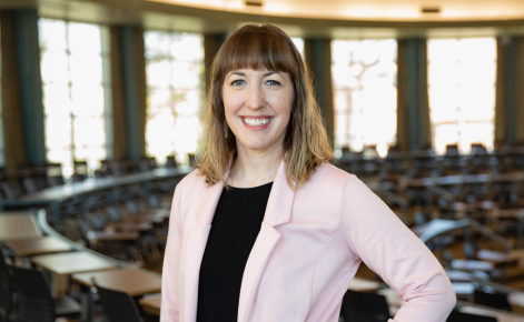 A person smiling and posing in a classroom setting, wearing a light pink blazer over a black top. The room features large windows and rows of desks and chairs.