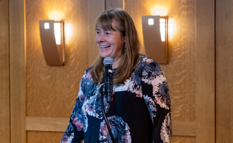 Karin Pfeiffer picture. Karin stands in front of a microphone and smiles. Karin wears a dark floral dress. Karin's blonde hair falls past the shoulders.