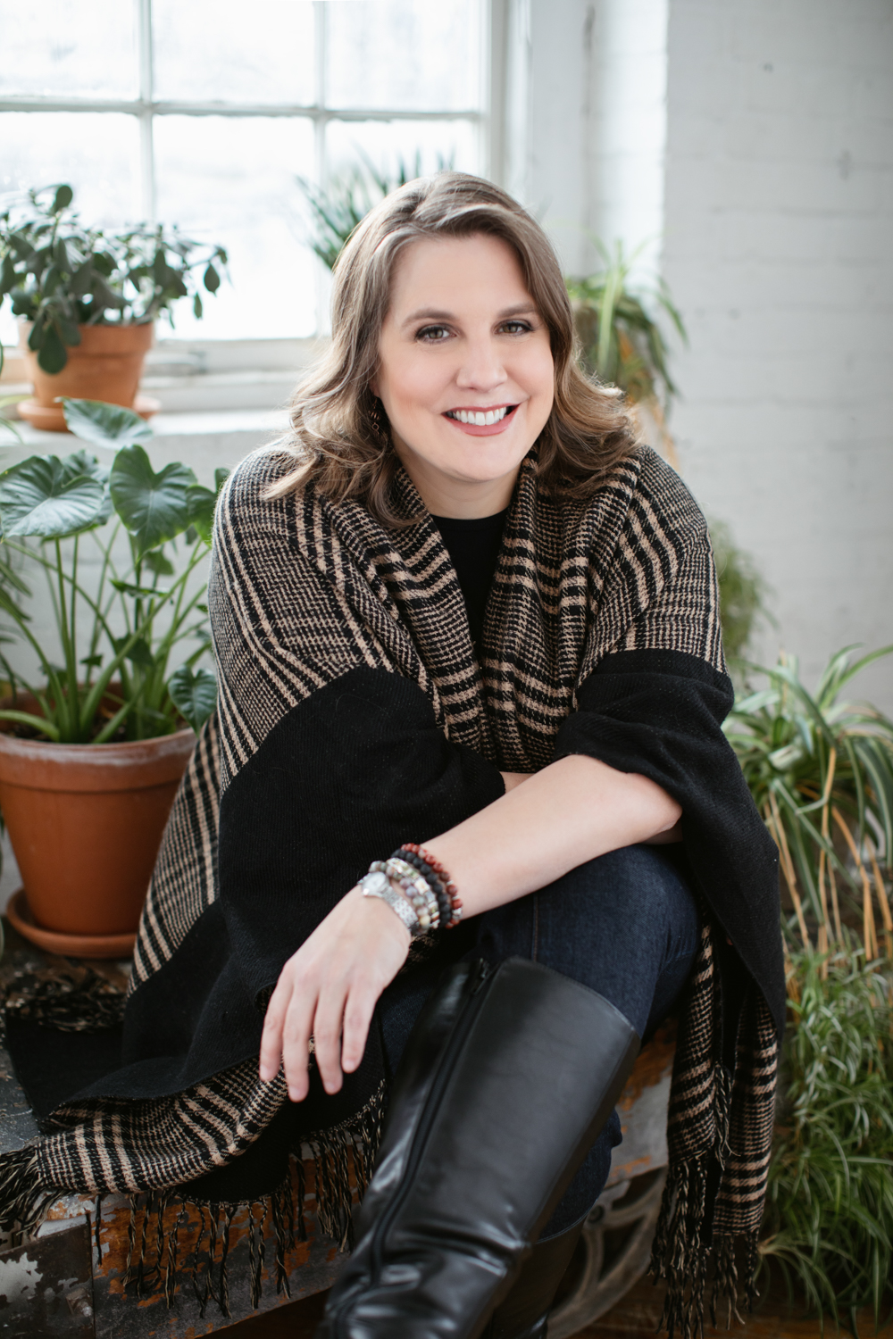 Shannon Burton headshot. Buton sits surrounded by potted plants and in front of a window. Shannon wears a black and tan shawl over a black shirt. Shannon wears bracelets of several colors.