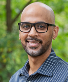 Flennaugh headshot. He wears a dark blue button down shirt with white polka dots on it. He has on dark-rimmed glasses.
