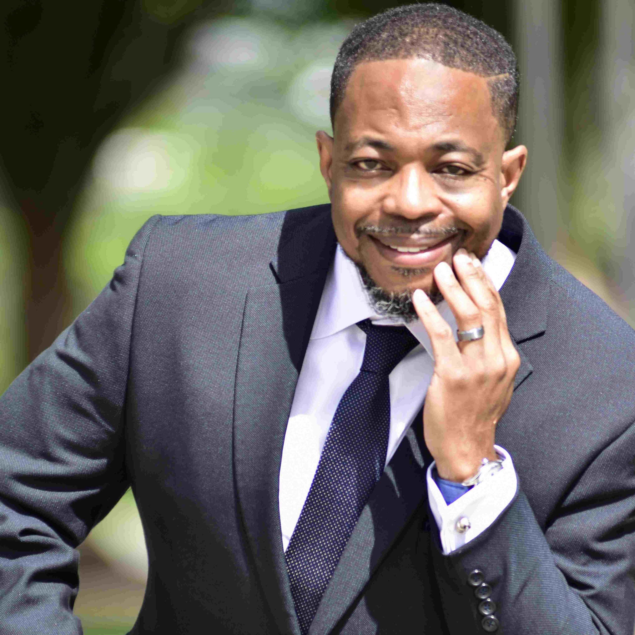 Ted Ransaw sits outside and smiles for the camera while bringing his hand up toward his face. Ransaw wears a white button-down shirt, a black tie and a black blazer. Ransaw has short black hair and a goatee.