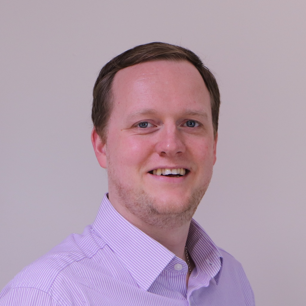 Headshot of Ian Cowburn. Ian is wearing a light purple button-down shirt. He stands against a neutral beige background. His brown hair is short.