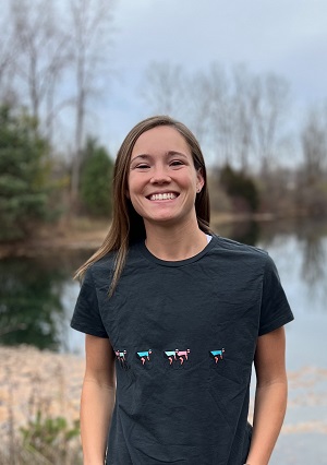 Corinne headshot. Corinne stands outside nearby a lake on an early spring day. Corinne wears a black t-shirt. Corinne has long brown hair.
