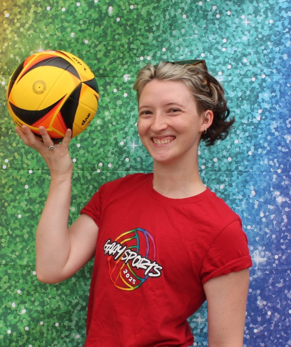 C. Davis smiles for the camera while holding up a yellow and black volleyball. Davis stands in front of a rainbow-gradient, sparkly background. Davis wears a red tee-shirt, and silver jewelry (rings, earrings). Davis' blonde hair is short, about chin-length.