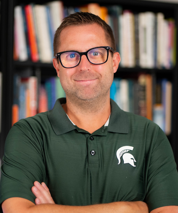 Scott Farver smiles for the camera in front of a full bookshelf. Farver wears an MSU-green polo shirt and dark-rimmed glasses. Farver has blonde hair cut close to the head.