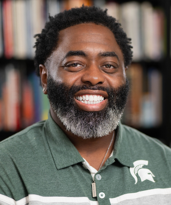 Curtis Lewis wearing a green Spartan polo. He is standing in front of a bookshelf. 