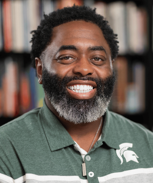 Curtis Lewis wearing a green Spartan polo. He is standing in front of a bookshelf. 