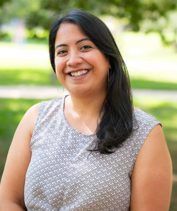 Noreen Naseem Rodríguez smiles for the camera in an outdoor setting. Rodríguez wears a sleeveless dress with a black and white lined pattern and hooped earrings in the shape of a pentagon. Rodríguez has black hair about shoulder-length.