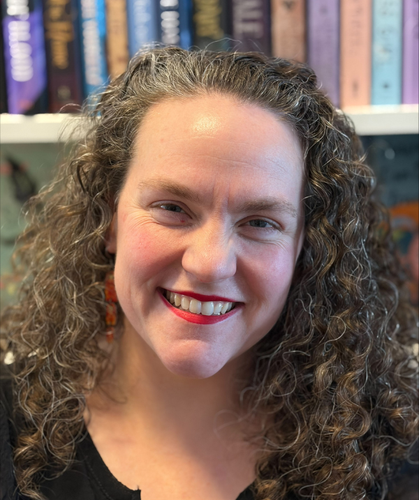 Cassie Sheets-Kavanagh smiles for the camera in front of a full bookshelf. Cassie wears a black top. Cassie's blonde hair is curly and falls past the shoulders.