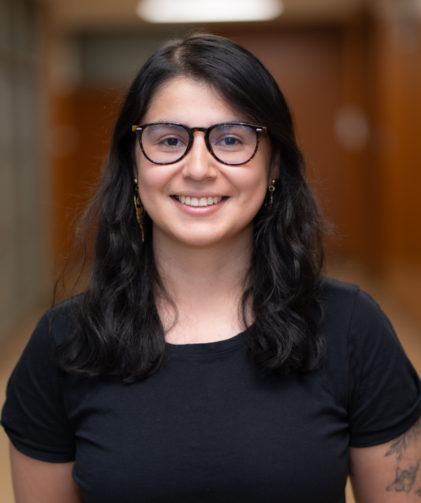 Angie Valbuena Rojas smiles for the camera while standing in a hallway in Erickson Hall. Angie wears a black top and black-rimmed glasses. Angie's hair is black, about shoulder-length, and wavy.