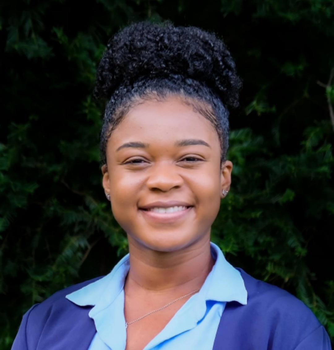 Chelsi Ricketts smiles for the camera in an outdoor setting, standing in front of an evergreen bush. Chelsi wears a blue top accented with a lighter blue collar and silver earrings. Chelsi's black hair is in a large bun on top of Chelsi's head.