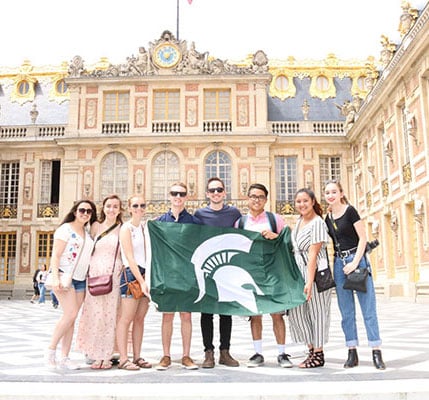 MSU Engineering student group holding a flag with the spartan helmet on it, standing in front of a building at CEA Paris Center