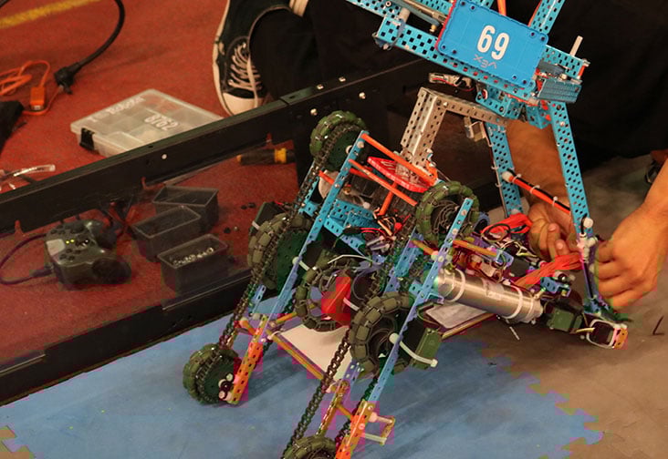 A kid bends over, focused on working on a VEX robotic machine car, with their face not visible. The image captures the child's concentration and engagement in building and programming the robotic vehicle.