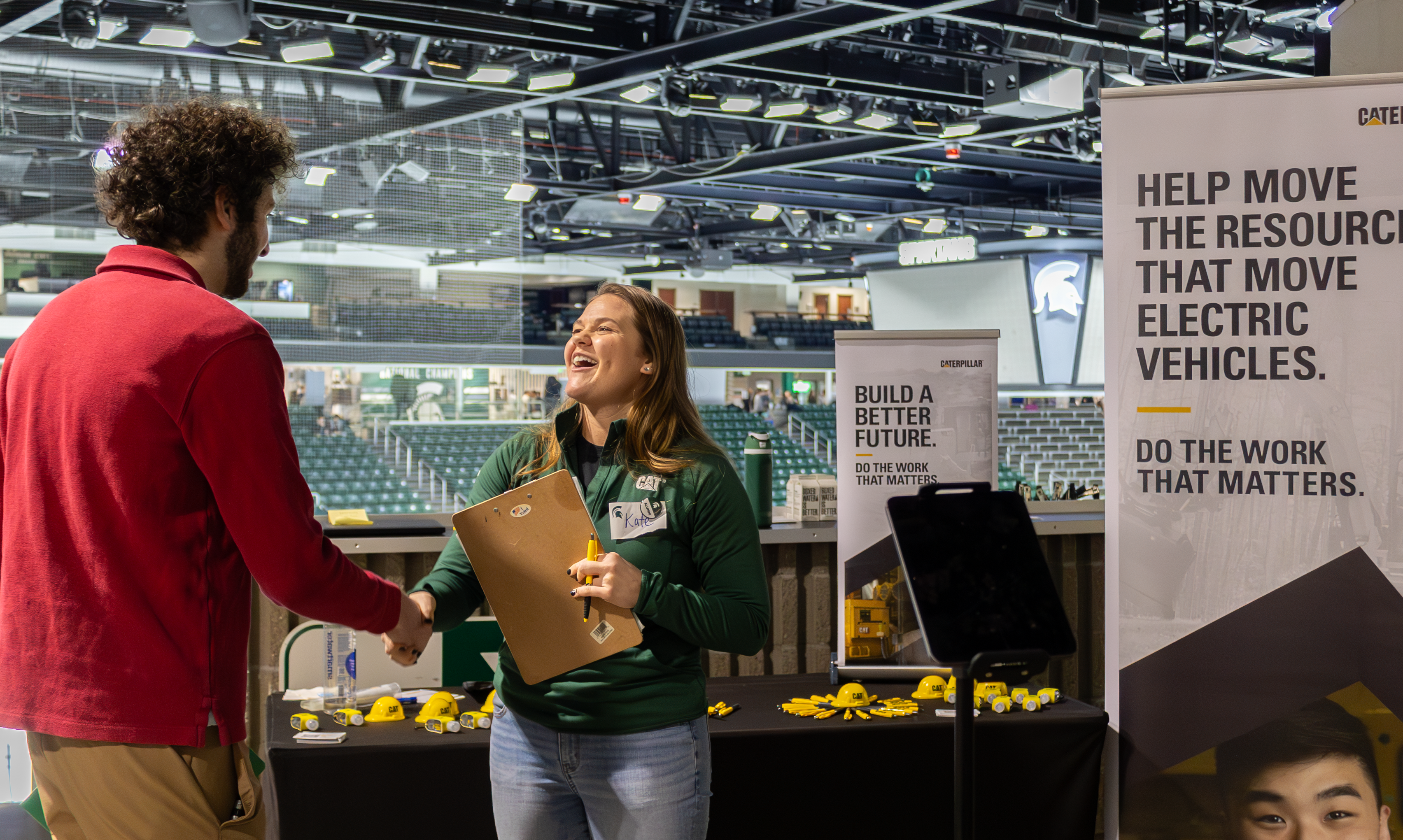 A recruiter at a booth laughs while shaking hands with an attendee at a career fair.
