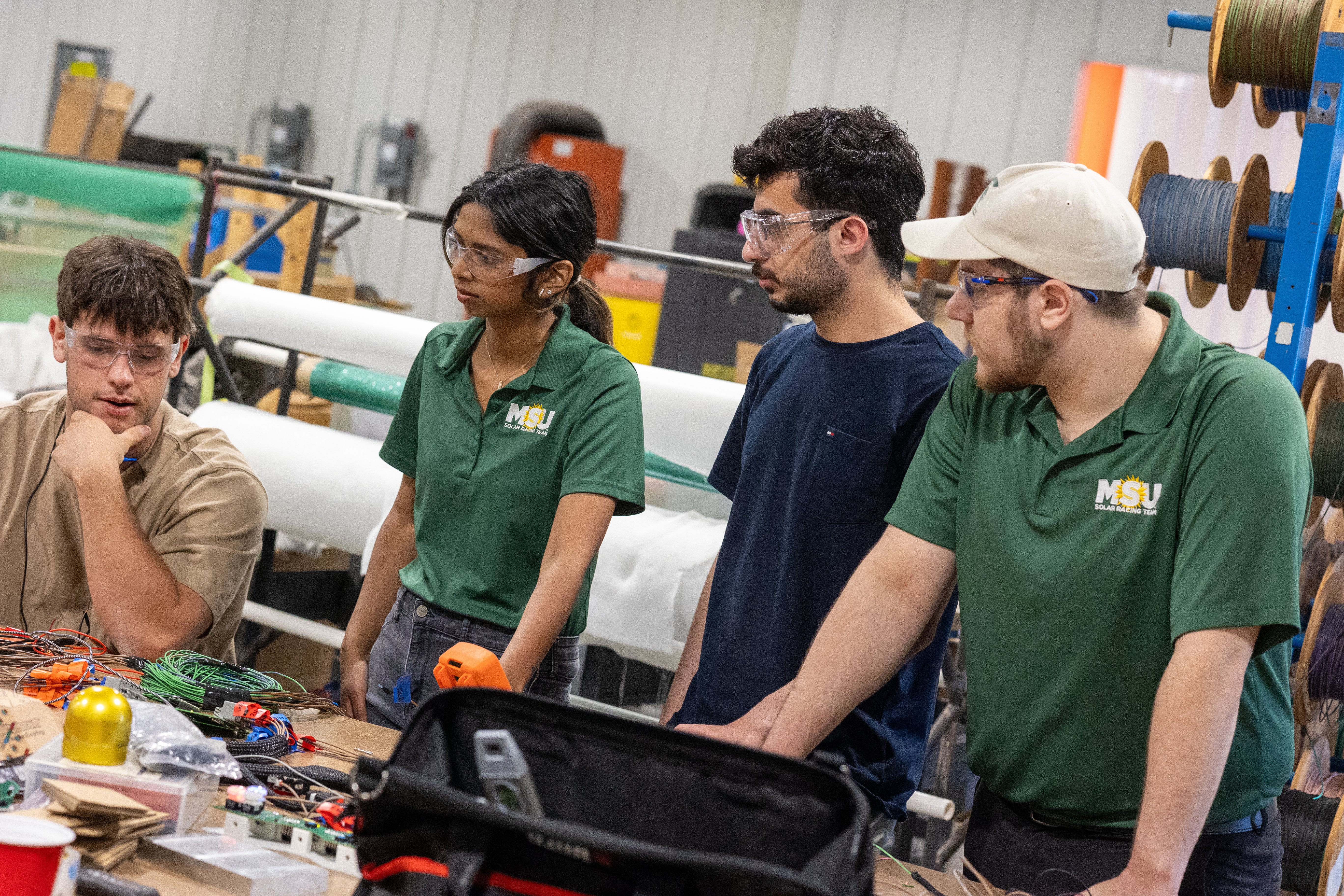 Four students wearing safety glasses collaborate on an engineering project with wires and tools.
