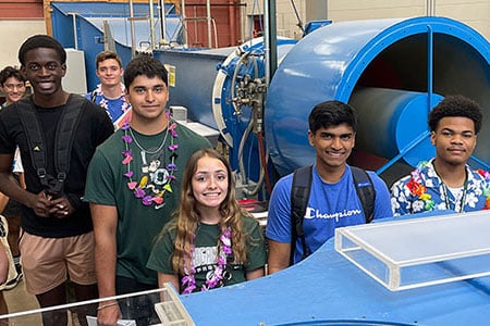 A group of students pose together in a lab with large blue wind tunnel equipment.