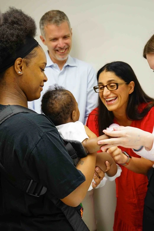 A photo with several people; one is holding a small baby while other people look on smiling.