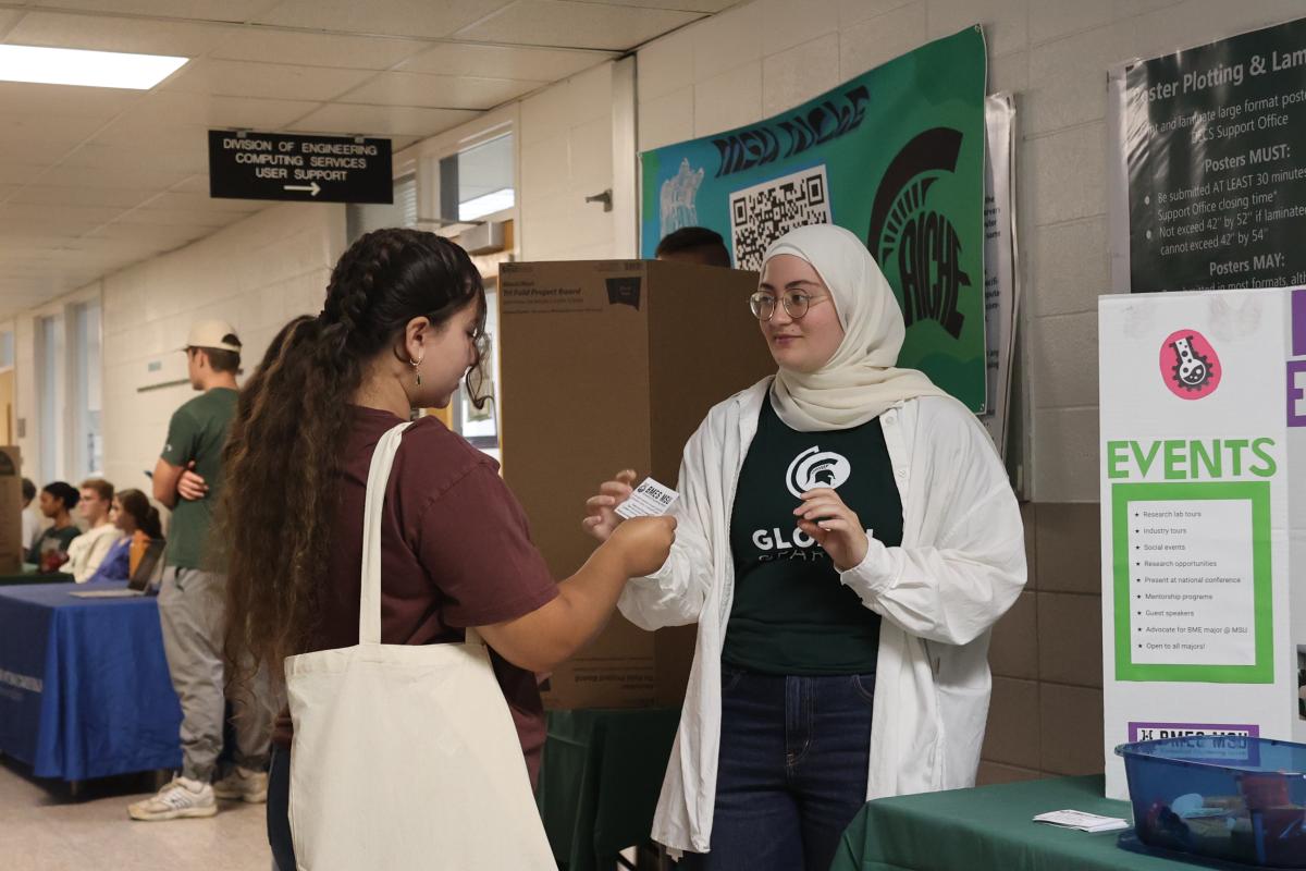 An individual wearing a white hijab, white oversized blouse and a green t-shirt with a spartan logo on it, hands a paper to another person with dark hair in a pony tail, wearing a maroon colored t-shirt and carrying a white canvas bag.