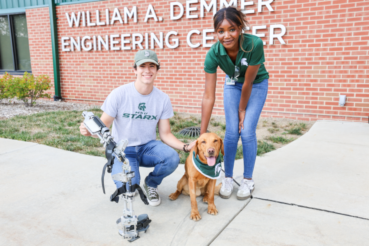 Two people petting a dog in front of a brick building with the sign that reads
