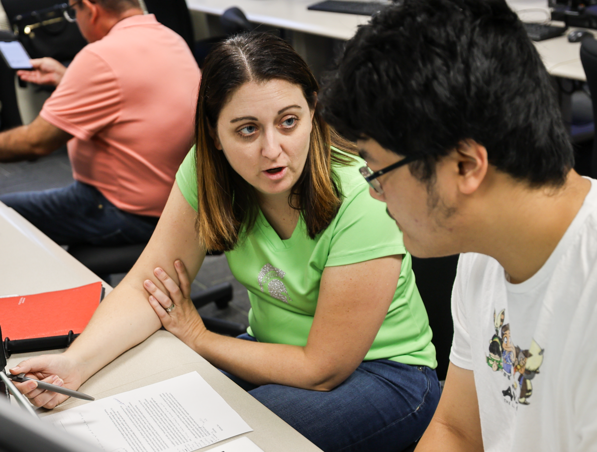 Woman with dark hair and a green shirt talking to a man with dark hair and glasses, wearing a white t-shirt.