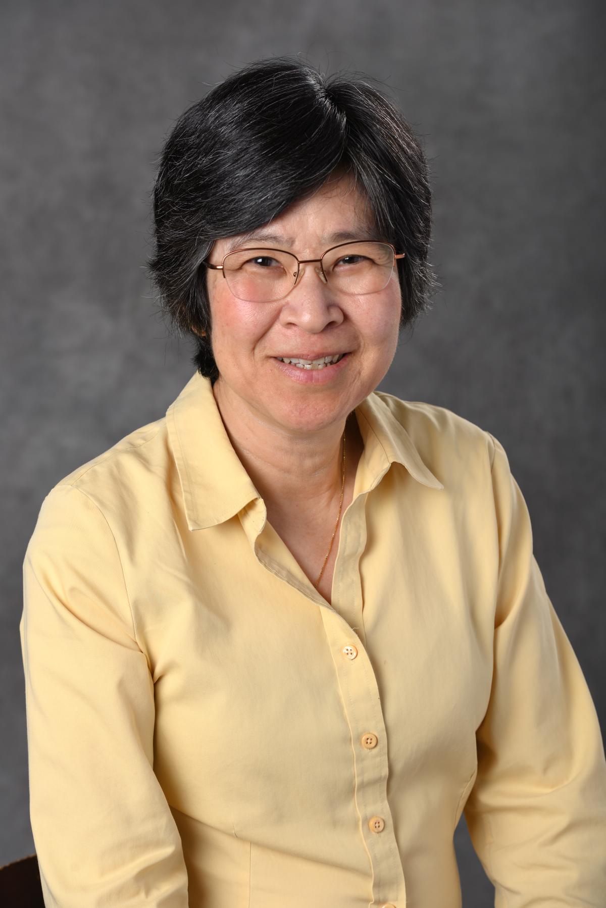 Person with dark hair and glasses wearing a yellow shirt, smiling, in front of a grey background.