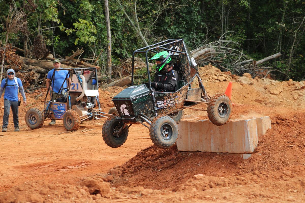 Baja-style vehicle navigating a raised barrier on a muddy course with people watching nearby.