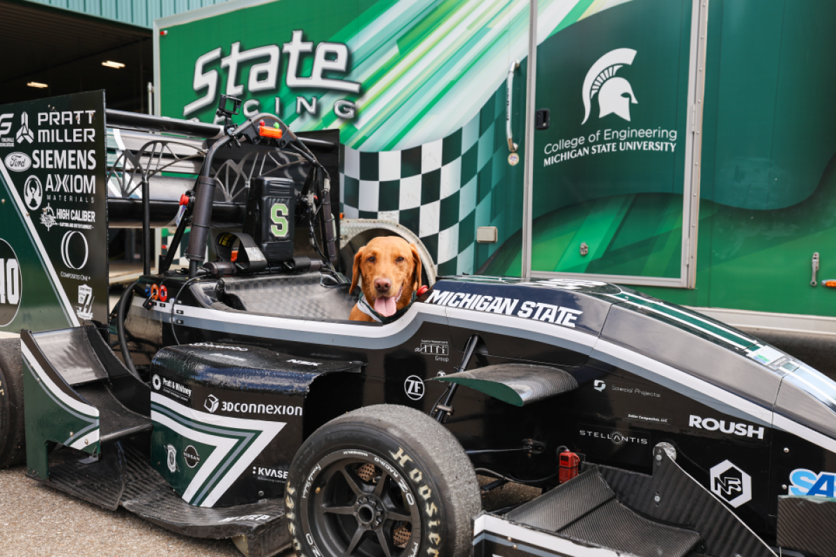 Zeke sitting in the cockpit of a Michigan State–branded race car with a green trailer in the background.