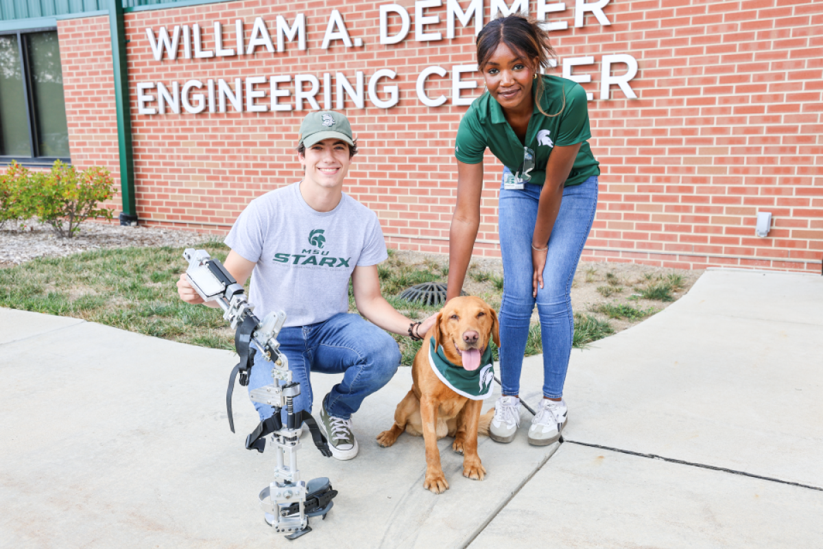 Two people posing with Zeke the Wonderdog wearing a bandana in front of a building