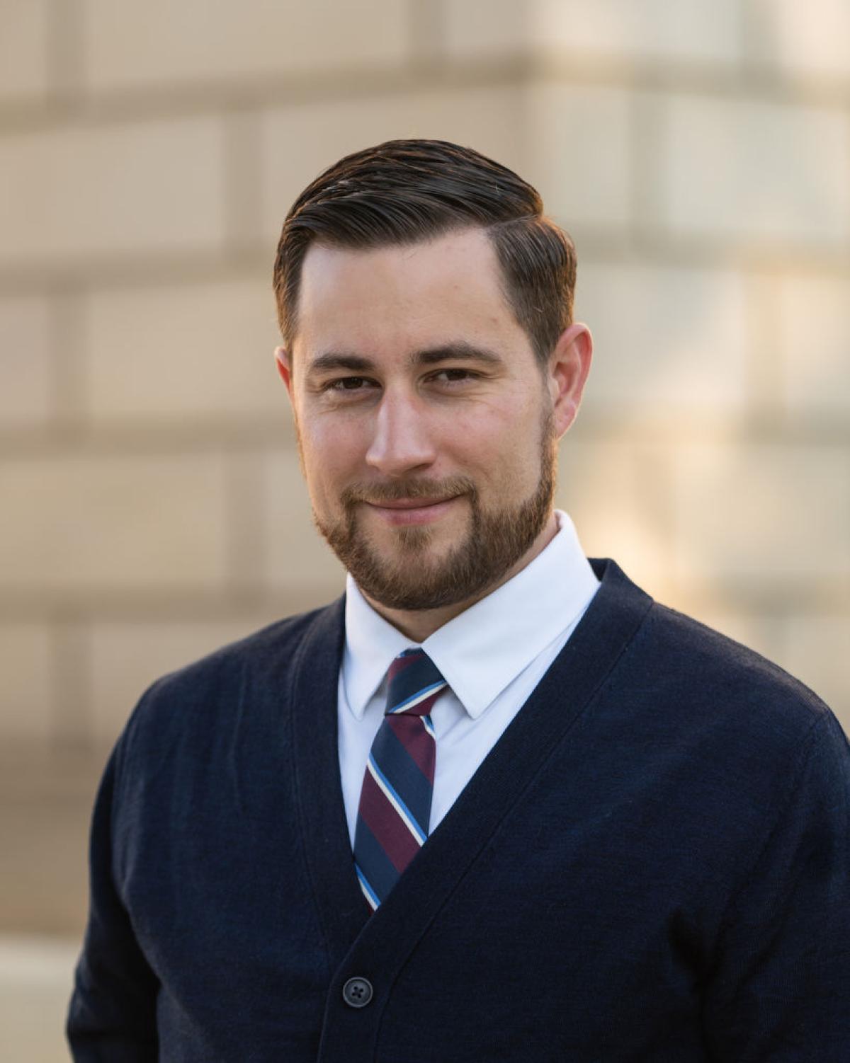 Judd Herzer wearing a dark sweater, white shirt and dark tie, smiling in front of a brick wall.