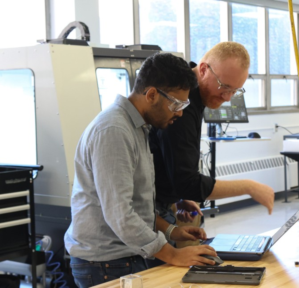 Two people stand at a workbench, focusing on a laptop, in a lab or engineering workspace.