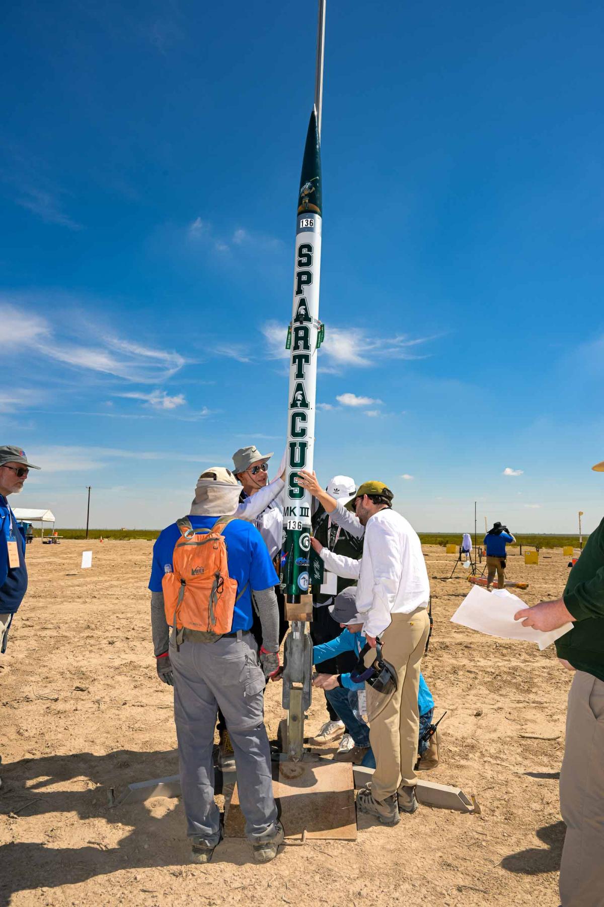  Several people adjusting a vertical rocket under a clear sky at a launch site.