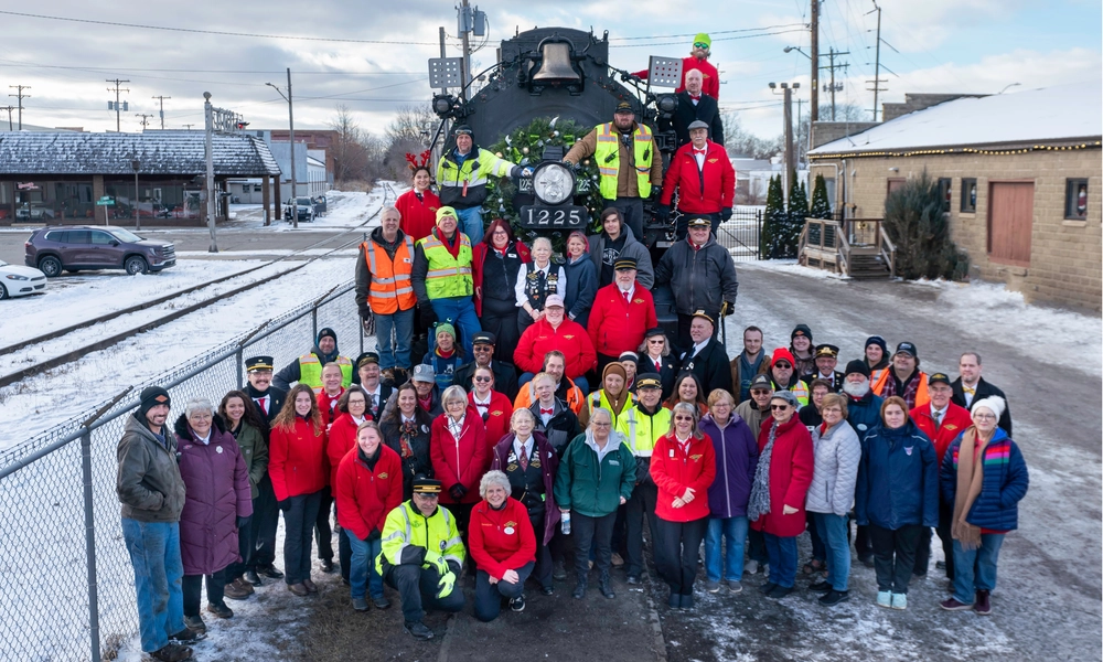 A team of crew members from the Steam Railroading Institute's North Pole Express stand smiling in front of the historic 1225 Pere Marquette steam engine.