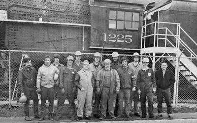 A group of men who are part of a rail crew, dressed in work clothes stands in front of a steam locomotive, smiling towards the camera.