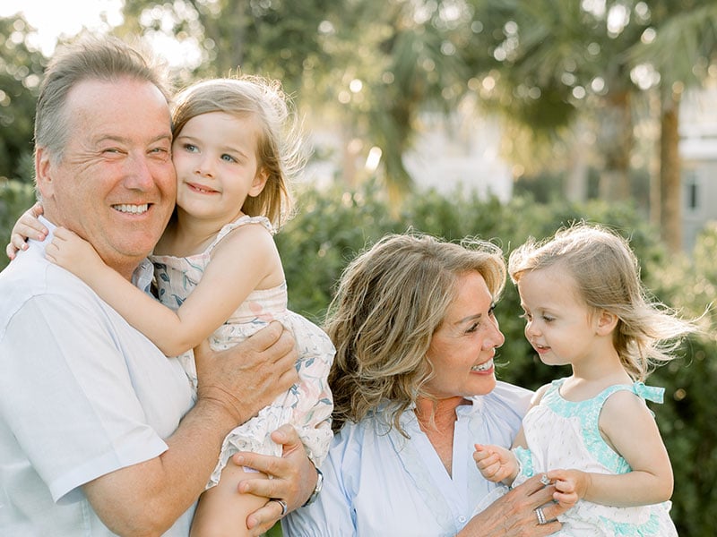 The Lamachs holding two young children outdoors with greenery and palm trees in the background.