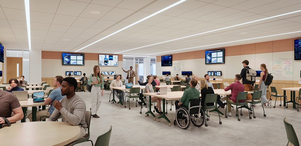 Collaborative classroom with round tables, laptops, and wall screens; instructor at front.