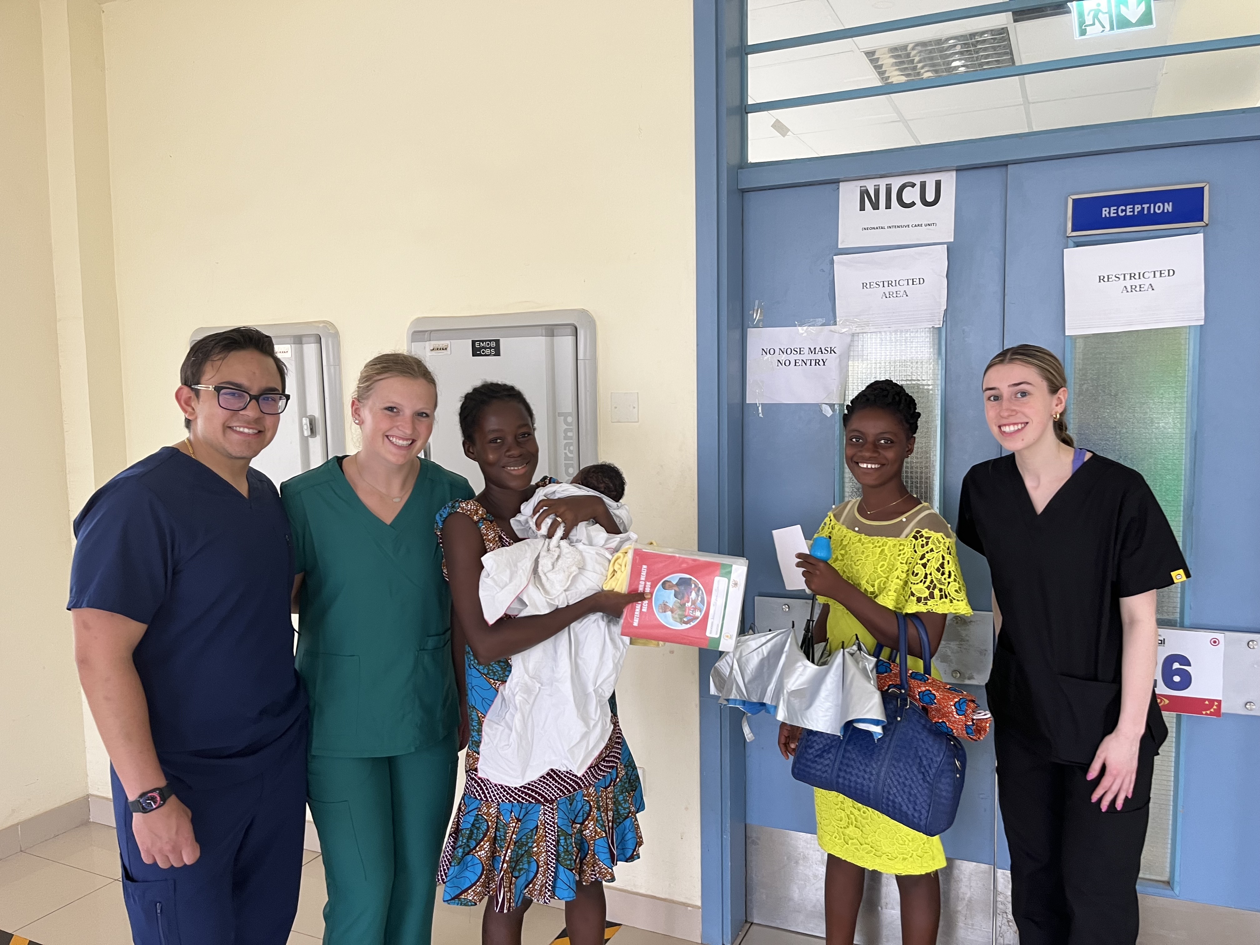 Three students in nursing uniforms stand with two women holding a baby.