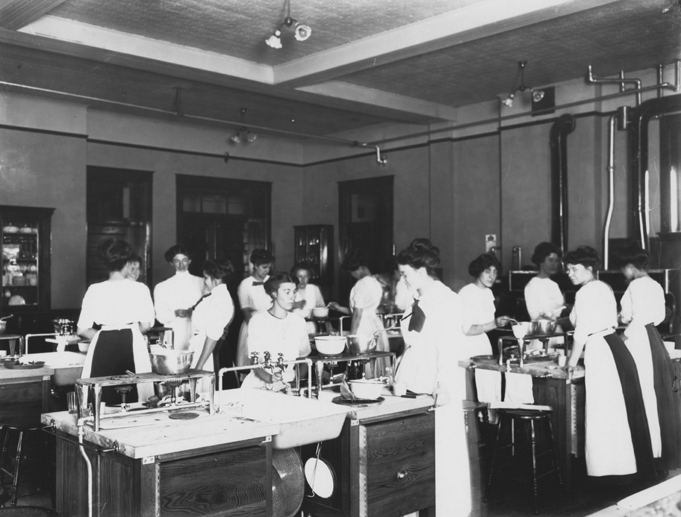 Women in long dresses in a classroom cooking.
