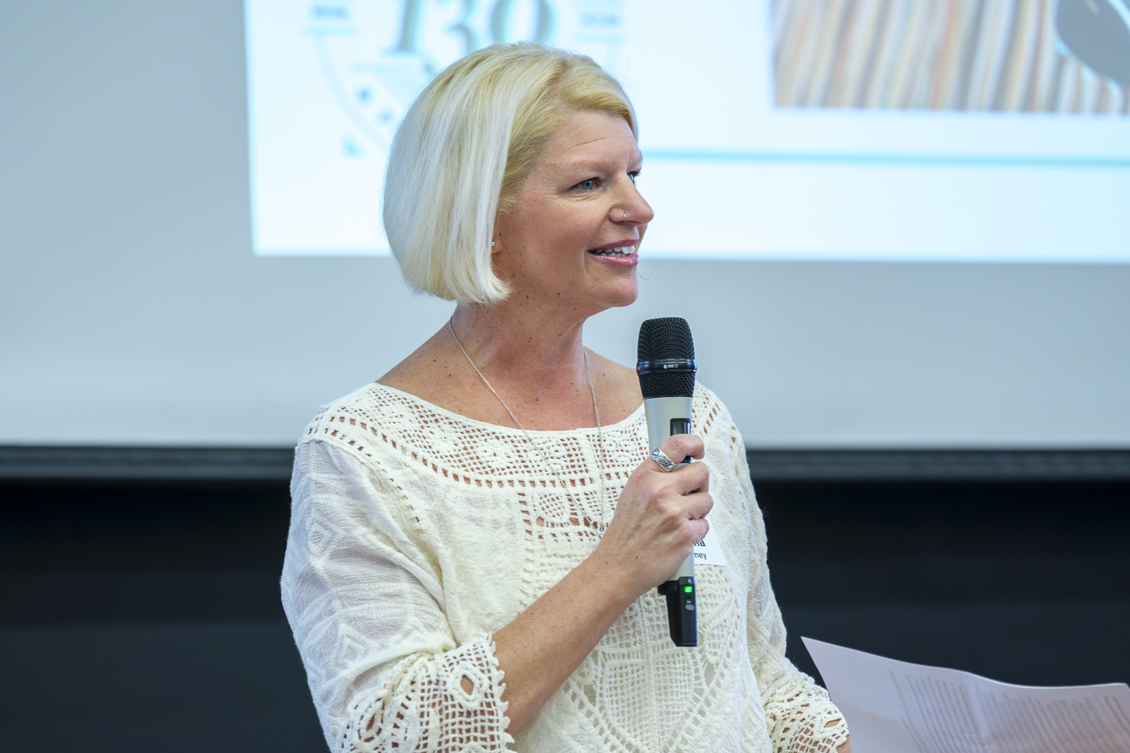 Woman speaks through microphone with a screen behind her.